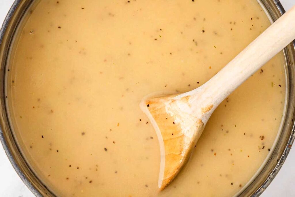 A wooden spoon stirs creamy, light brown gravy with black pepper in a saucepan, perfect for your Friendsgiving feast.