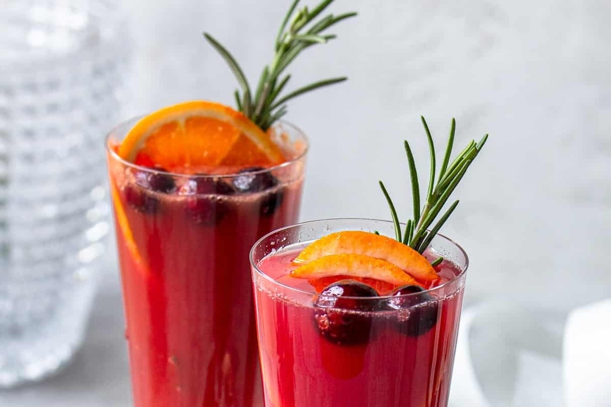 Two glasses of cranberry drinks garnished with orange slices and rosemary sprigs.