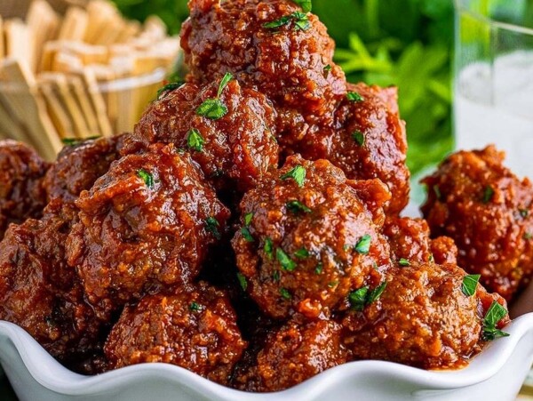 A white bowl filled with saucy meatballs—perfect as cranberry appetizers—garnished with herbs, with a glass of ice and toothpicks in the background.