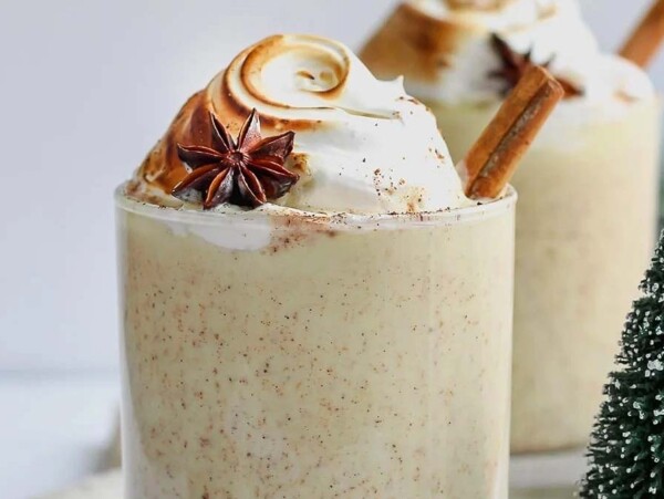 Glass of eggnog topped with whipped cream, star anise, and a cinnamon stick—perfect inspiration for your holiday eggnog drinks recipes, with festive decorations nearby.