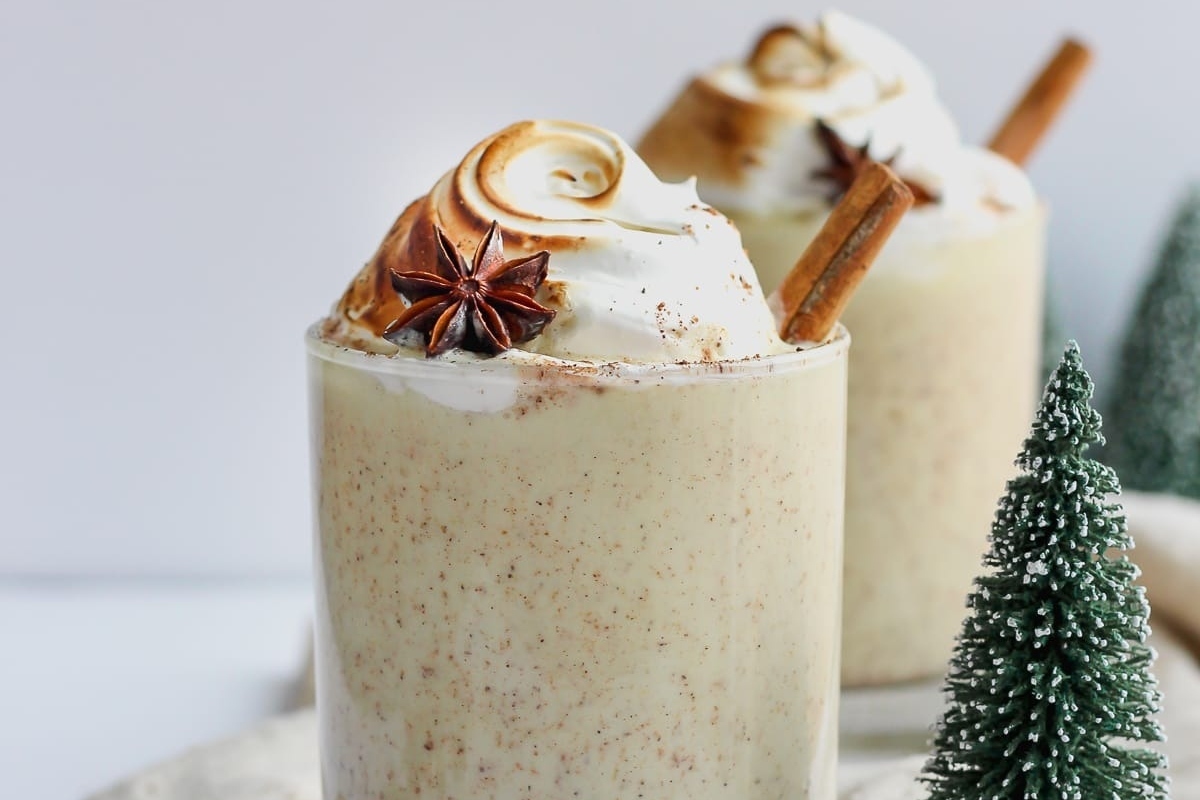 Two glasses of eggnog topped with whipped cream and cinnamon sticks.