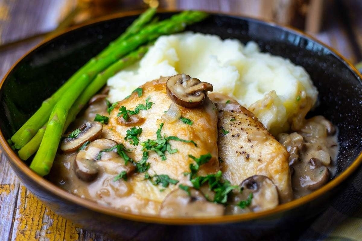 A plate of chicken with mushroom sauce, mashed potatoes, and green asparagus, served in a black bowl on a wooden table.