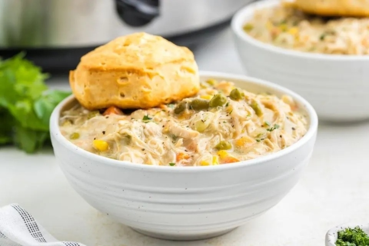 A bowl of creamy chicken soup with vegetables and a biscuit on top, served on a white countertop.