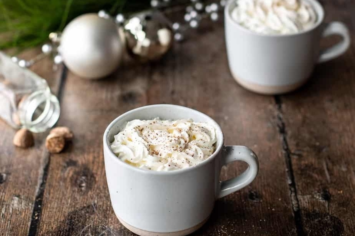 Two mugs of hot cocoa with whipped cream on a wooden table, perfect for indulging in cozy eggnog-inspired drinks recipes.