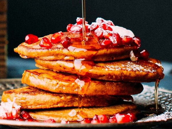 A stack of pancakes topped with pomegranate seeds, cream, and syrup being poured over them—one of the festive christmas breakfast recipes perfect for a joyful morning.