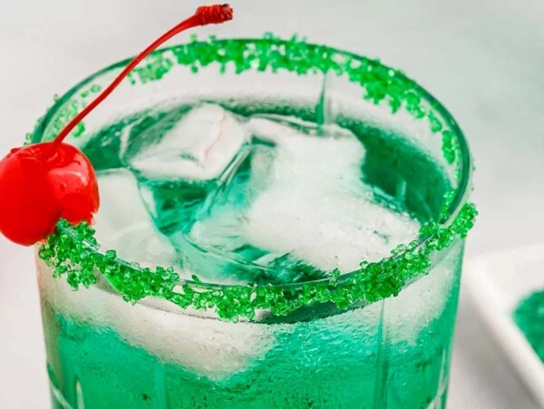 Close-up of a green Christmas drink with ice, a green sugar rim, and a cherry garnish on the edge of the glass.