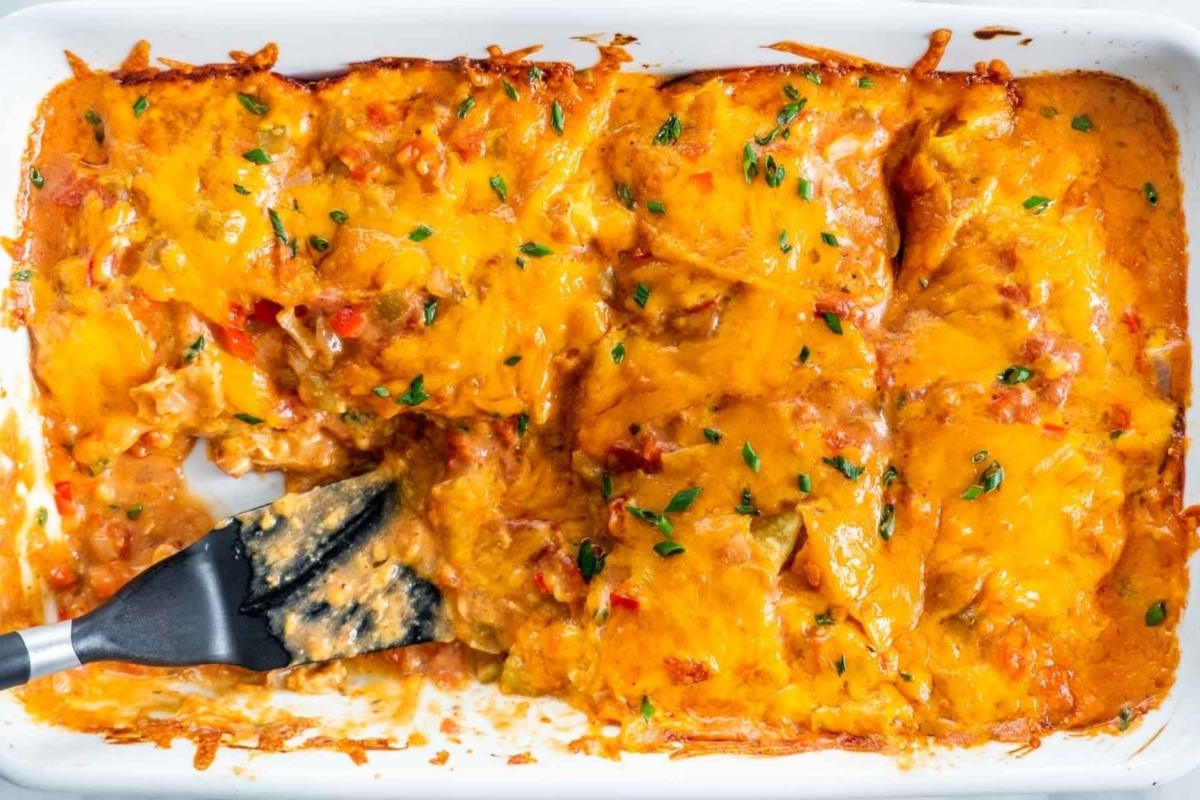 Baked enchiladas covered in melted cheese and garnished with herbs in a white dish, with a serving spatula.