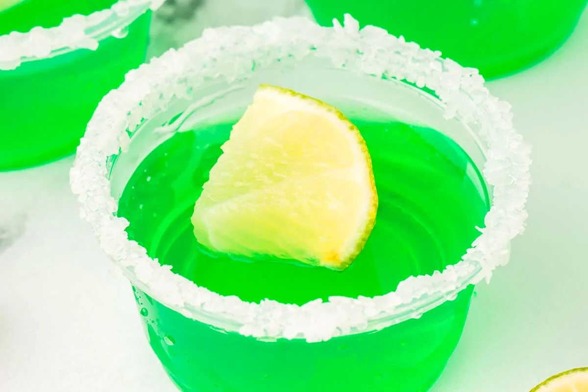 A lime margarita in a glass with a slice of lime and jello shots.