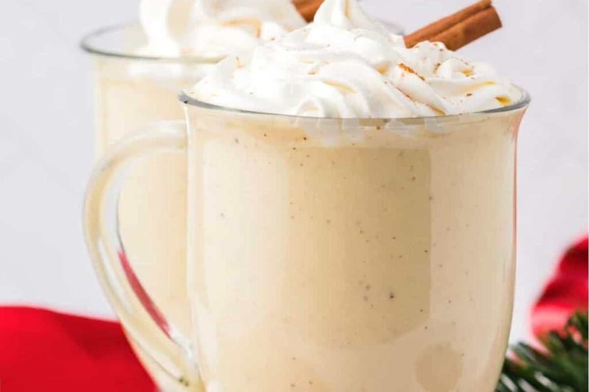 Two eggnog mugs with whipped cream and cinnamon sticks.