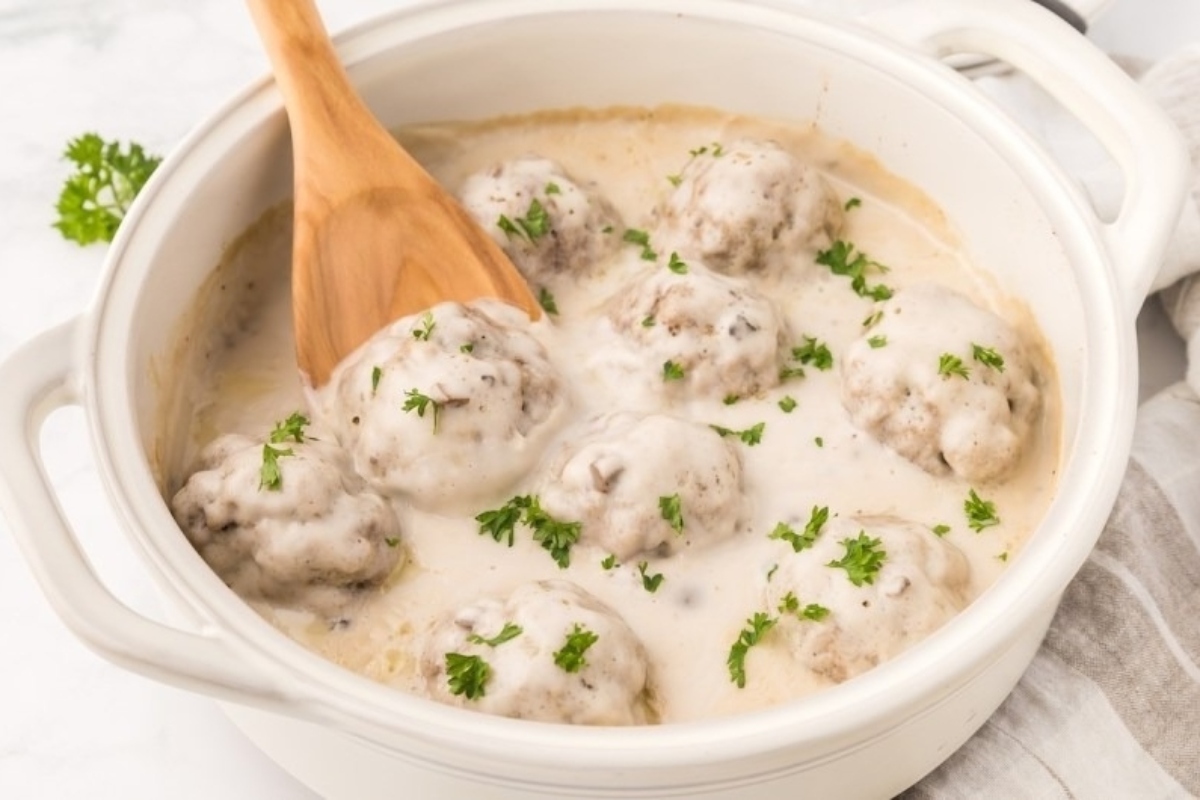 Swedish meatballs in a creamy sauce garnished with parsley in a white casserole dish with a wooden spoon.