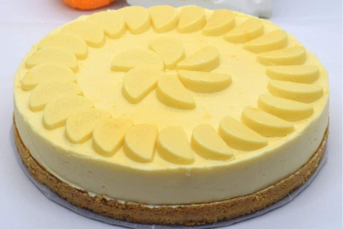 A festive yellow cheesecake, perfect for a no bake Christmas dessert, is sitting on a table next to oranges.
