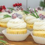 White Chocolate Cranberry Cupcakes with white frosting, sugared cranberries, and rosemary sprigs are beautifully arranged on a wooden tray.