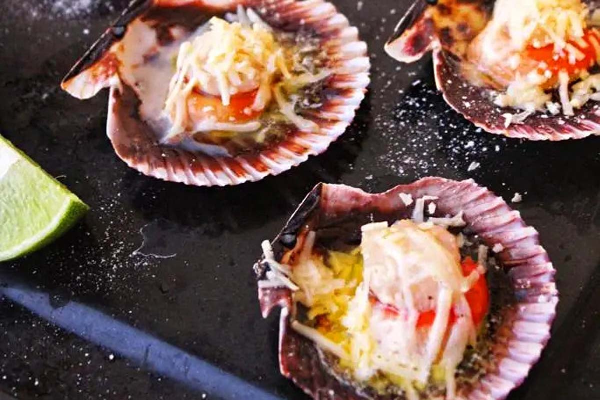 Four scallop shells with tomatoes and cheese on a black tray, prepared for the feast of seven fishes.