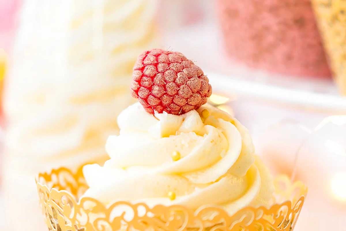 A cupcake with swirled yellow frosting topped with a frosted raspberry in a decorative gold wrapper—perfect for New Year's Eve desserts.