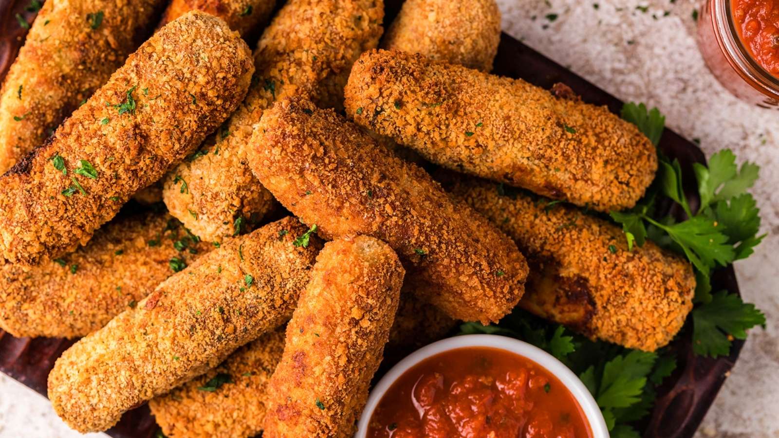 A pile of breaded, golden-brown mozzarella sticks—perfect for air fryer recipes—sits next to a bowl of marinara sauce, garnished with parsley.
