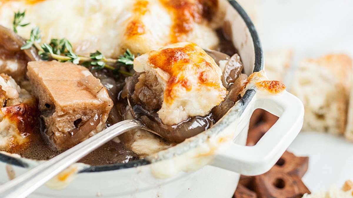 A bowl of French onion soup, one of the classic soup recipes, topped with melted cheese and bread, with a spoon inside.