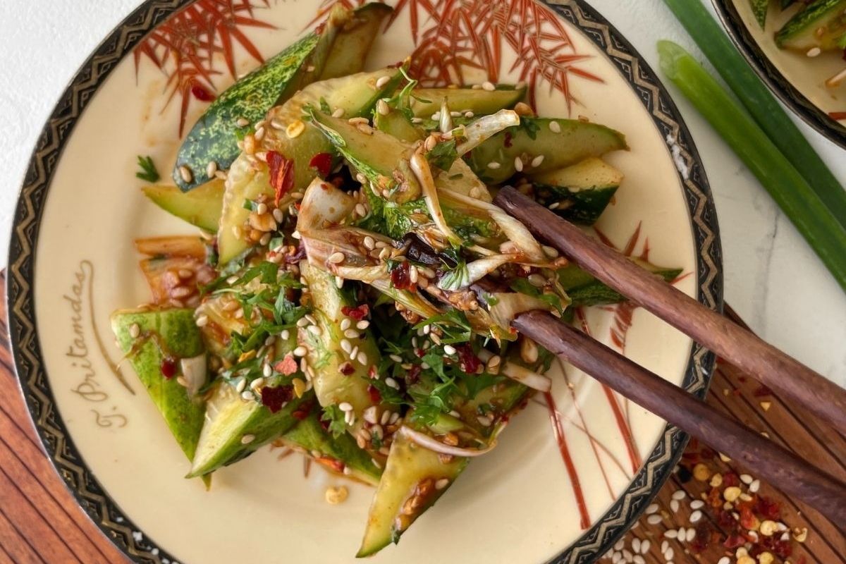 Chopsticks holding spicy cucumber salad with sesame seeds and herbs on a decorative plate—perfect for adding freshness to your Lunar New Year recipes.