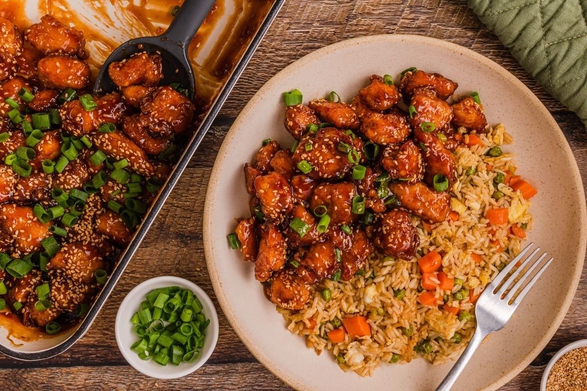 Plate of fried rice with vegetables and glazed chicken, topped with green onions and sesame seeds on a wooden table—a delicious addition to your collection of Lunar New Year recipes.