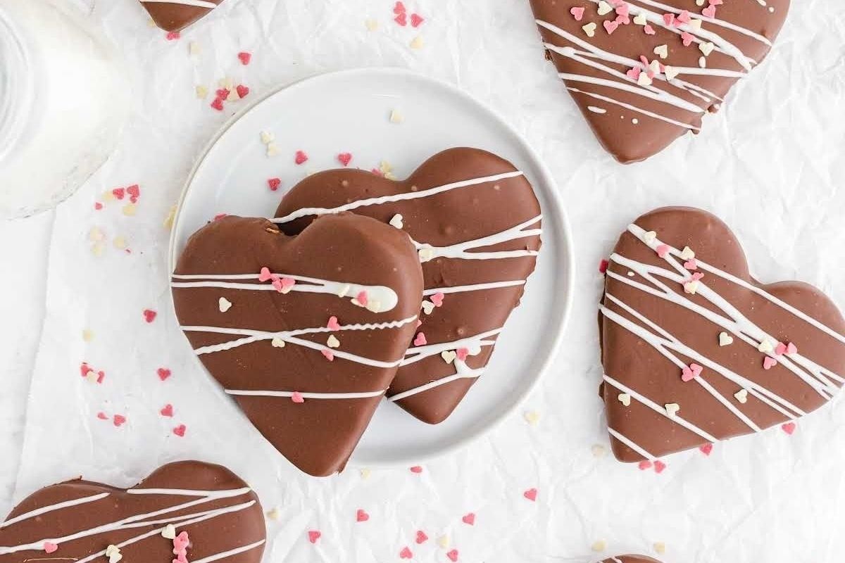 Cute Valentine's treats: Heart-shaped chocolate cookies with white drizzle and pink heart sprinkles, arranged on a plate and parchment paper.