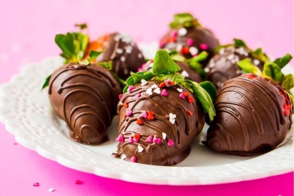 Cute Valentine's treats: chocolate-covered strawberries with colorful sprinkles on a white plate against a pink background.