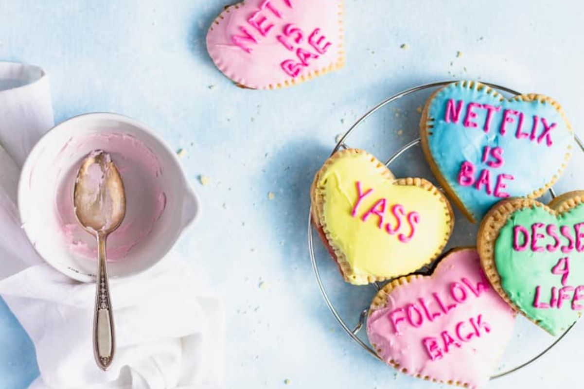 Heart-shaped cookies with colorful icing and playful phrases like "NETFLIX IS BAE" and "FOLLOW BACK"—a sweet idea for Valentine's breakfasts for kids, displayed on a cheerful blue surface.