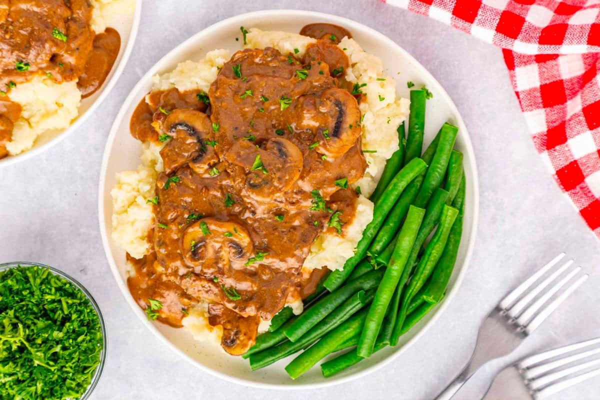 A plate of mashed potatoes topped with salisbury steak and mushroom gravy, served with a side of green beans, on a table with a red-checkered napkin.