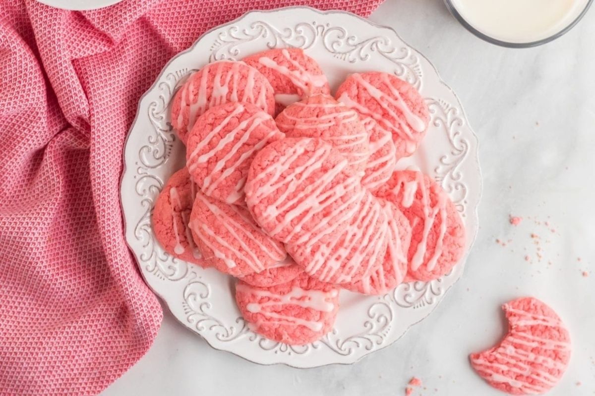 A plate of pink Valentine's Day cookies with white drizzle icing sits next to a pink cloth, with a bitten cookie on the side.
