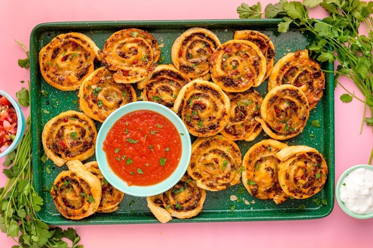A tray of freshly baked pinwheel pizza rolls with dipping sauces and garnish on a pink background.