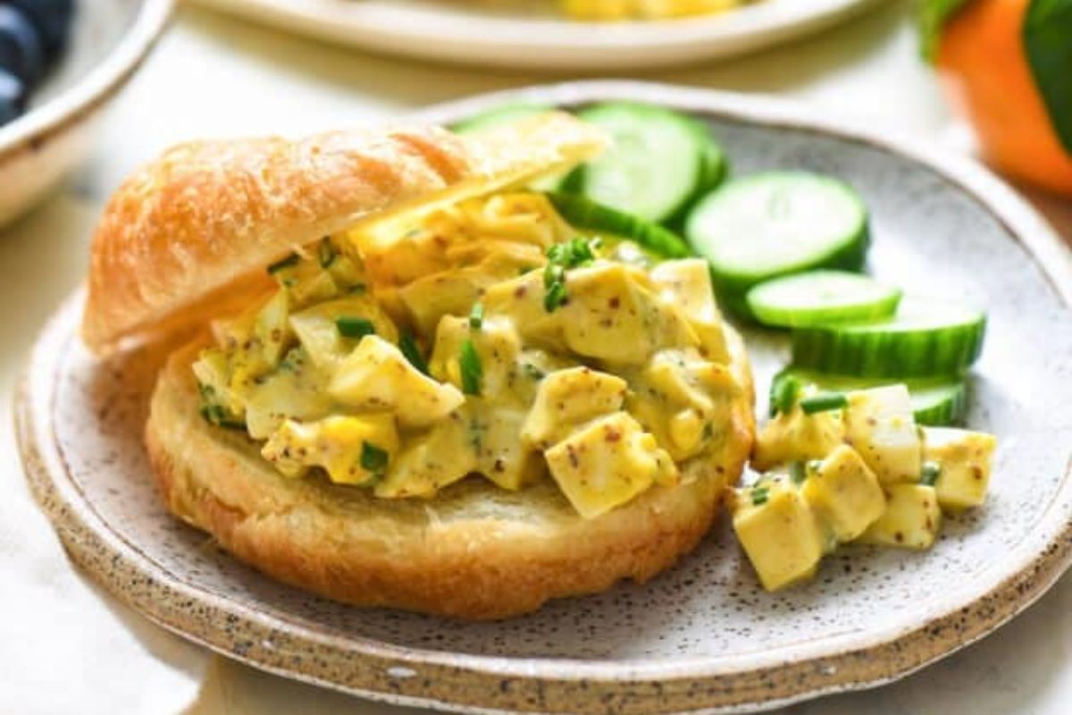 An egg salad sandwich on a plate with pickles and blueberries.