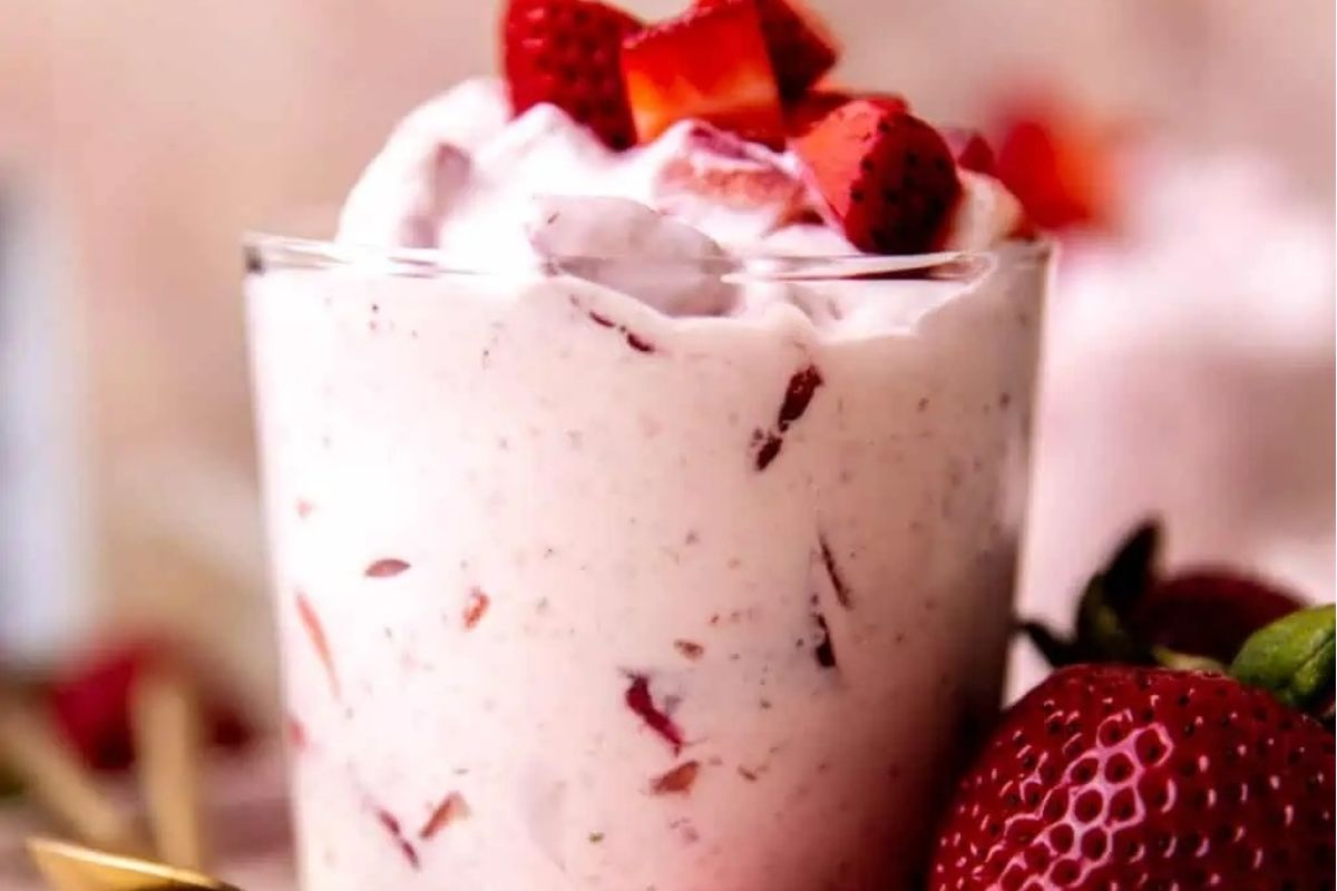 A pink strawberry smoothie in a glass with whipped cream and strawberries.