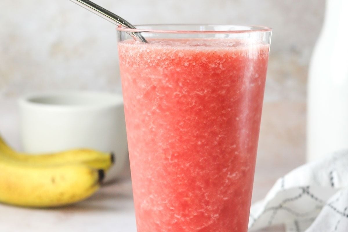 A glass of watermelon smoothie with a straw, bananas, and pinwheel recipes.