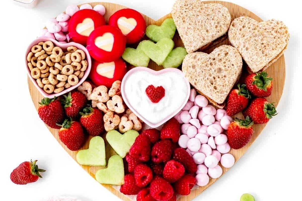 Heart-shaped board with strawberries, raspberries, crackers, cheese, bread, yogurt, and snacks arranged decoratively—perfect for Valentine's breakfasts for kids.