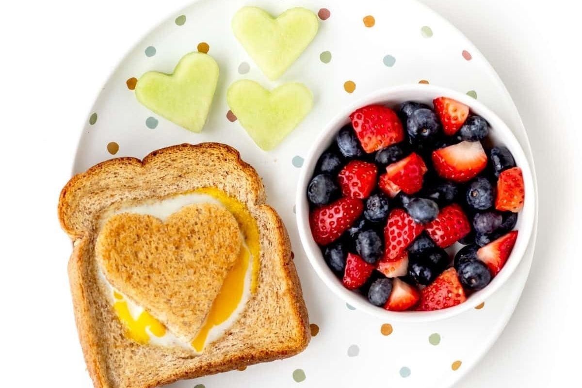 A plate with heart-shaped toast and egg, two melon hearts, and a bowl of strawberries and blueberries—perfect for Valentine's breakfasts for kids.