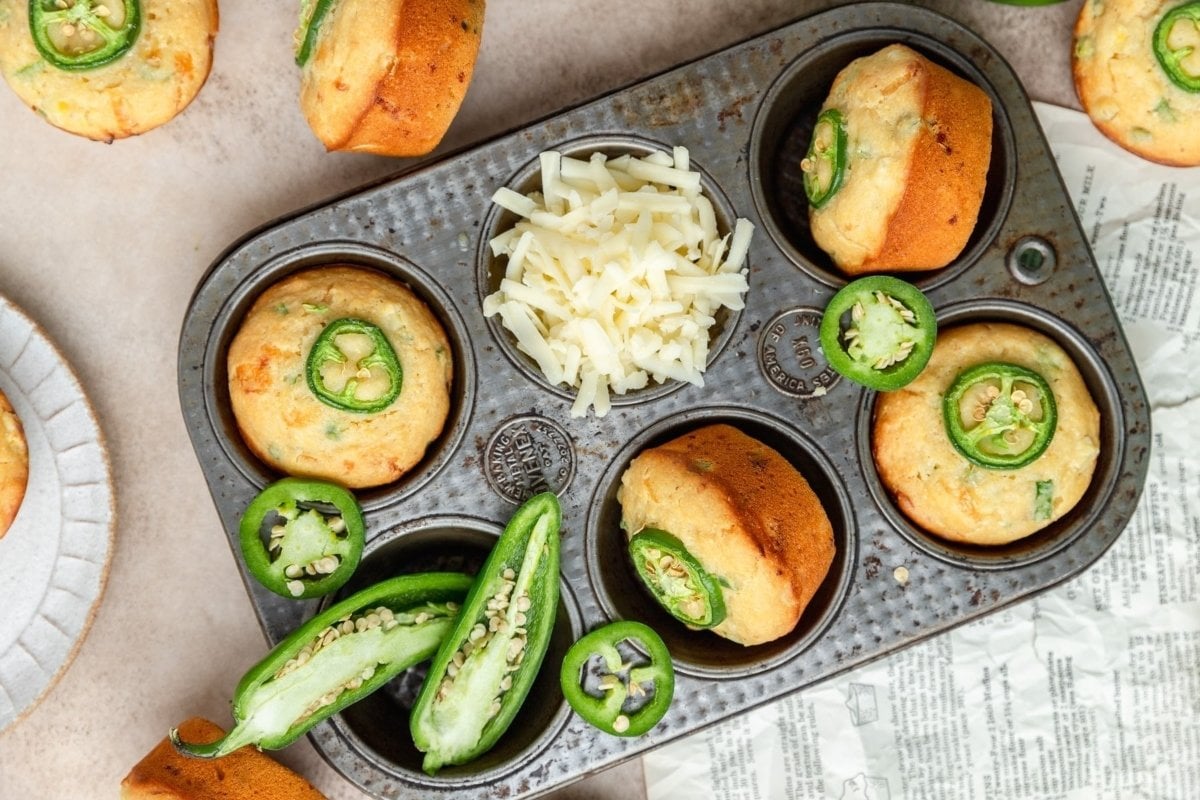 Freshly baked savory jalapeño muffins in a muffin tin, garnished with sliced jalapeños and shredded cheese, placed on a table.