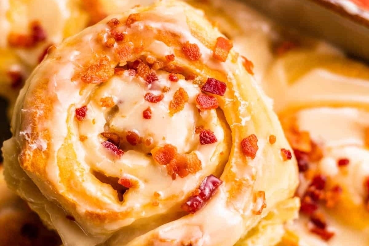 A mouthwatering close up of a cinnamon roll topped with crispy bacon and drizzled with sweet icing.