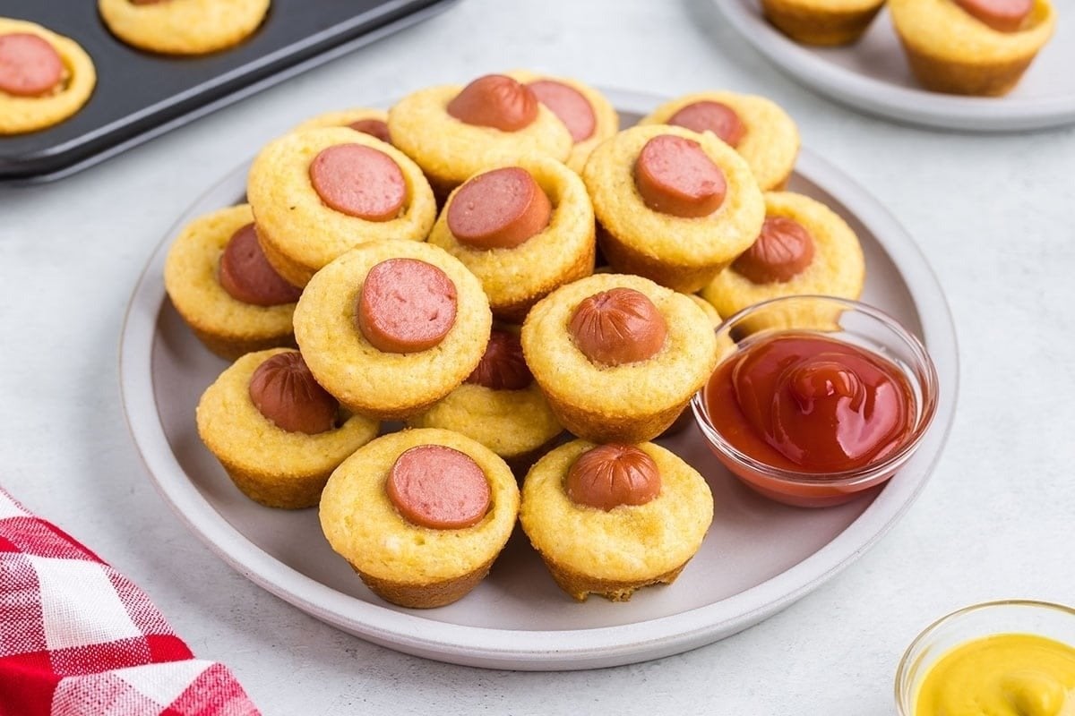A plate of savory cornbread muffins with hot dog slices on top, served with a side of ketchup.