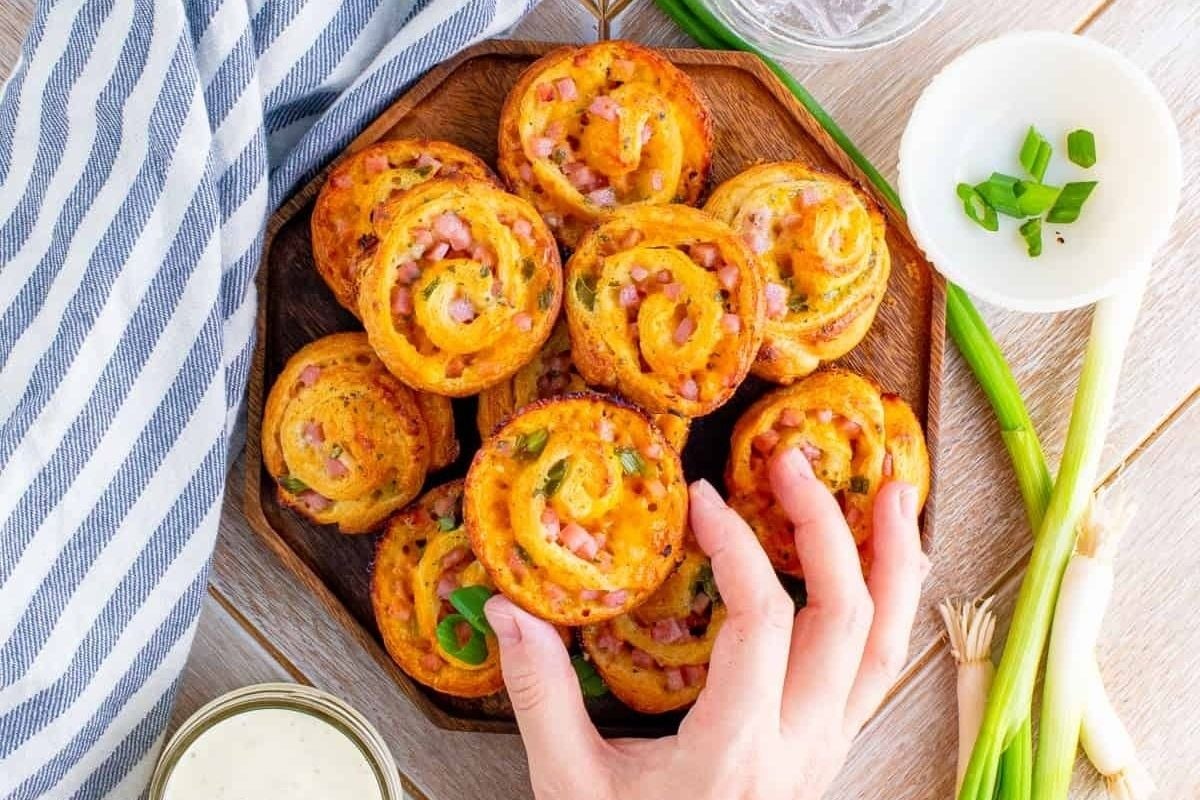 A hand picking up a pinwheel appetizer from a wooden platter garnished with green onions, showcasing one of the many delicious pinwheel recipes.