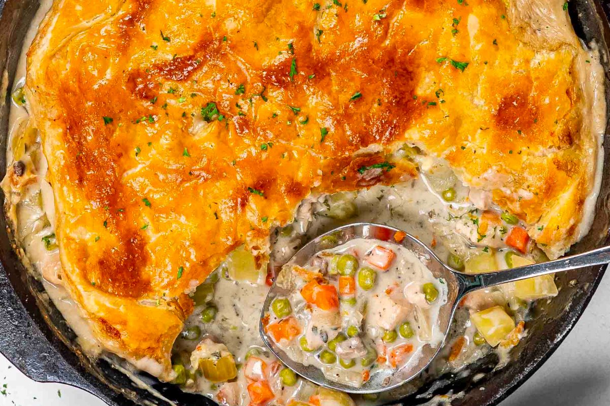 A skillet of creamy chicken pot pie with a golden-brown crust, partially scooped with a spoon.