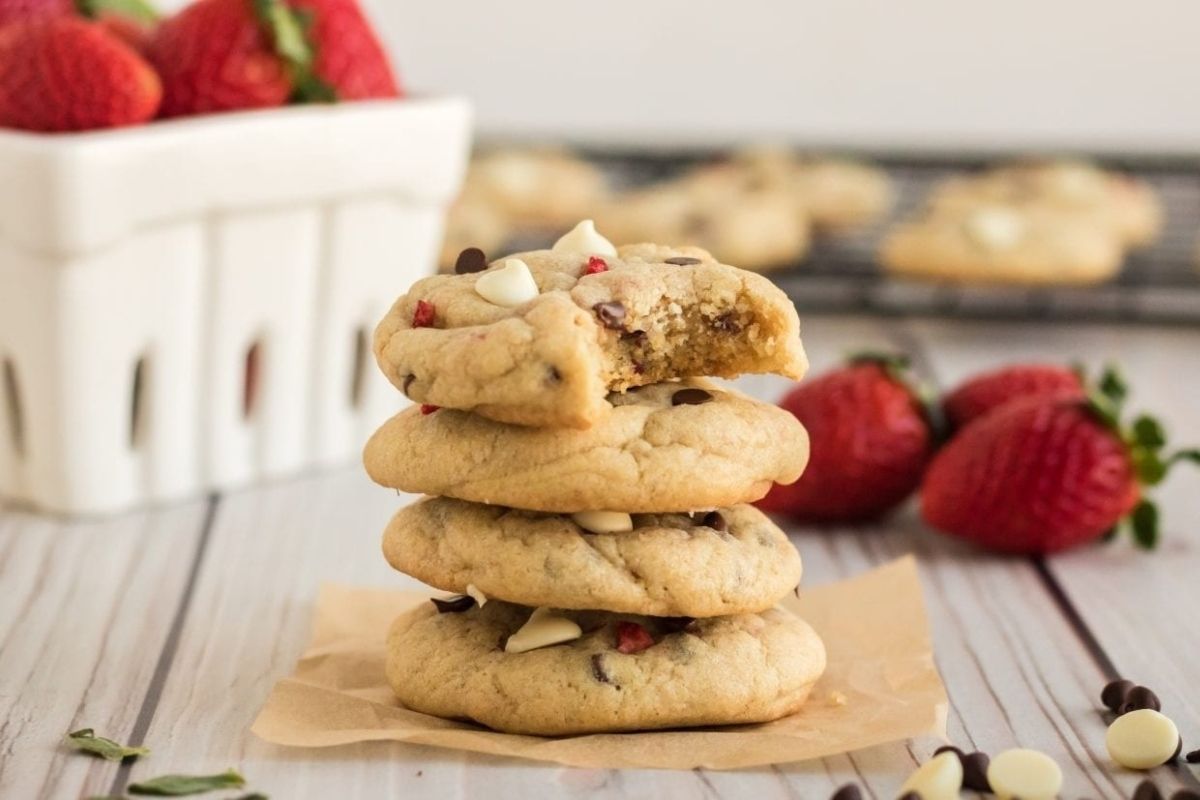 A stack of Valentine's Day cookies with chocolate chips and white chips, surrounded by strawberries, with a white basket of strawberries in the background.