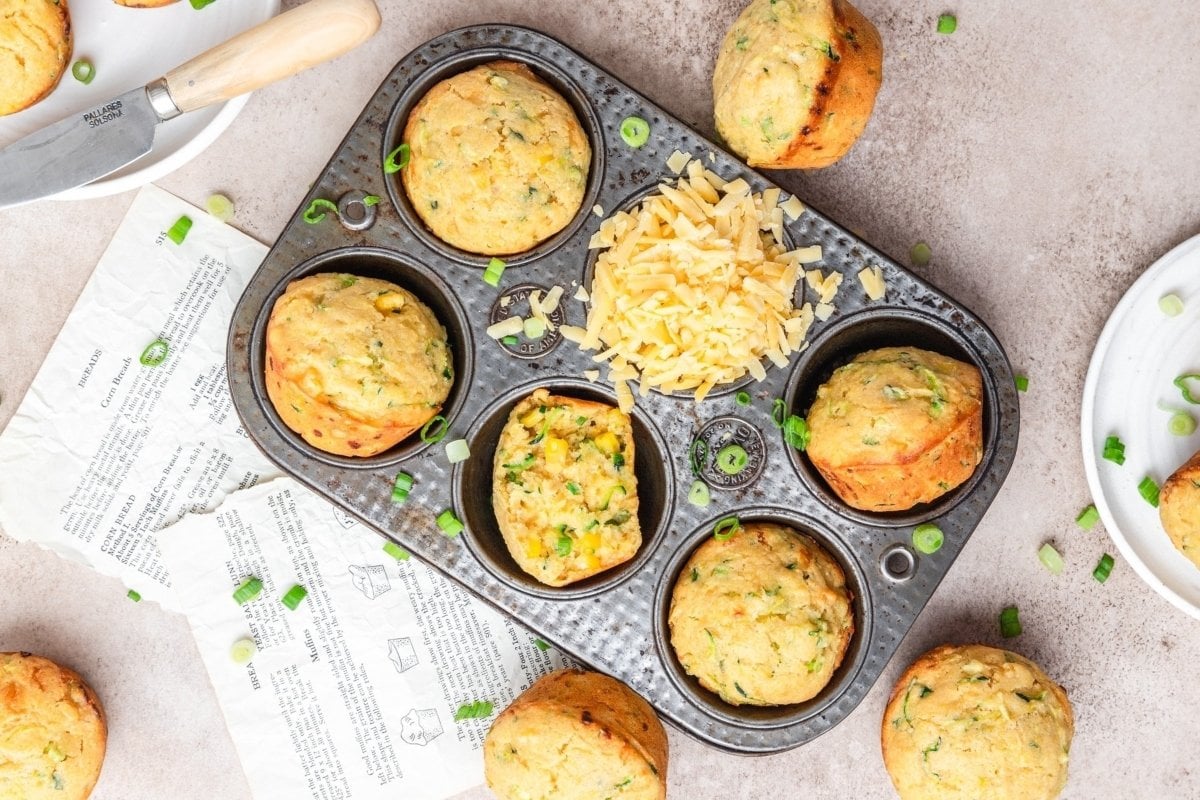 A platter of savory muffins just out of the oven, filled with cheese and green onions, some ingredients visible on the side.
