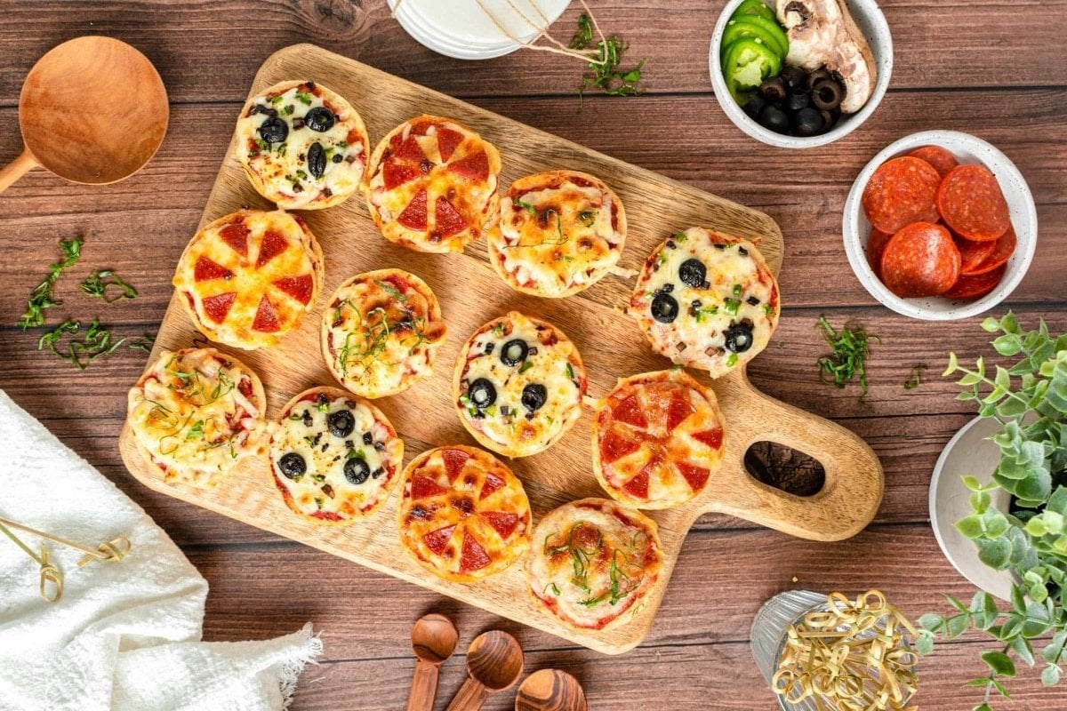 Assorted mini pizzas on a wooden board, bites for dinner, featuring toppings like olives, pepperoni, and vegetables, on a rustic table with herbs and cooking utensils.