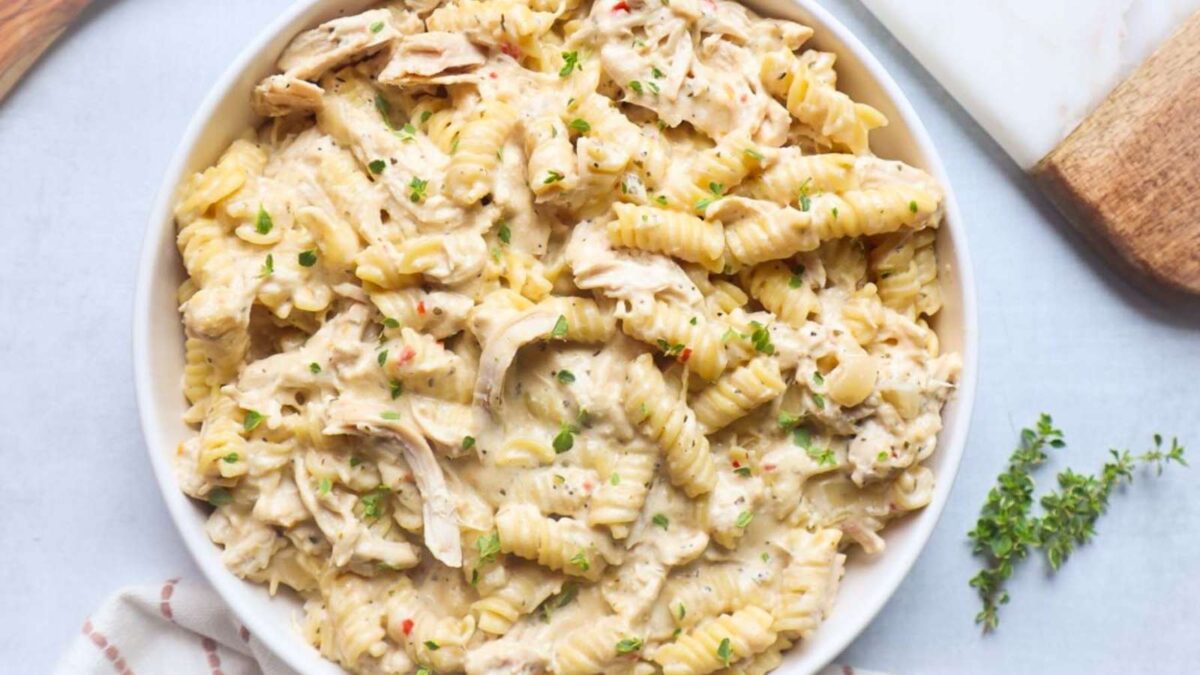 A bowl of creamy rotini pasta with shredded chicken, prepared in a slow cooker and garnished with chopped herbs.
