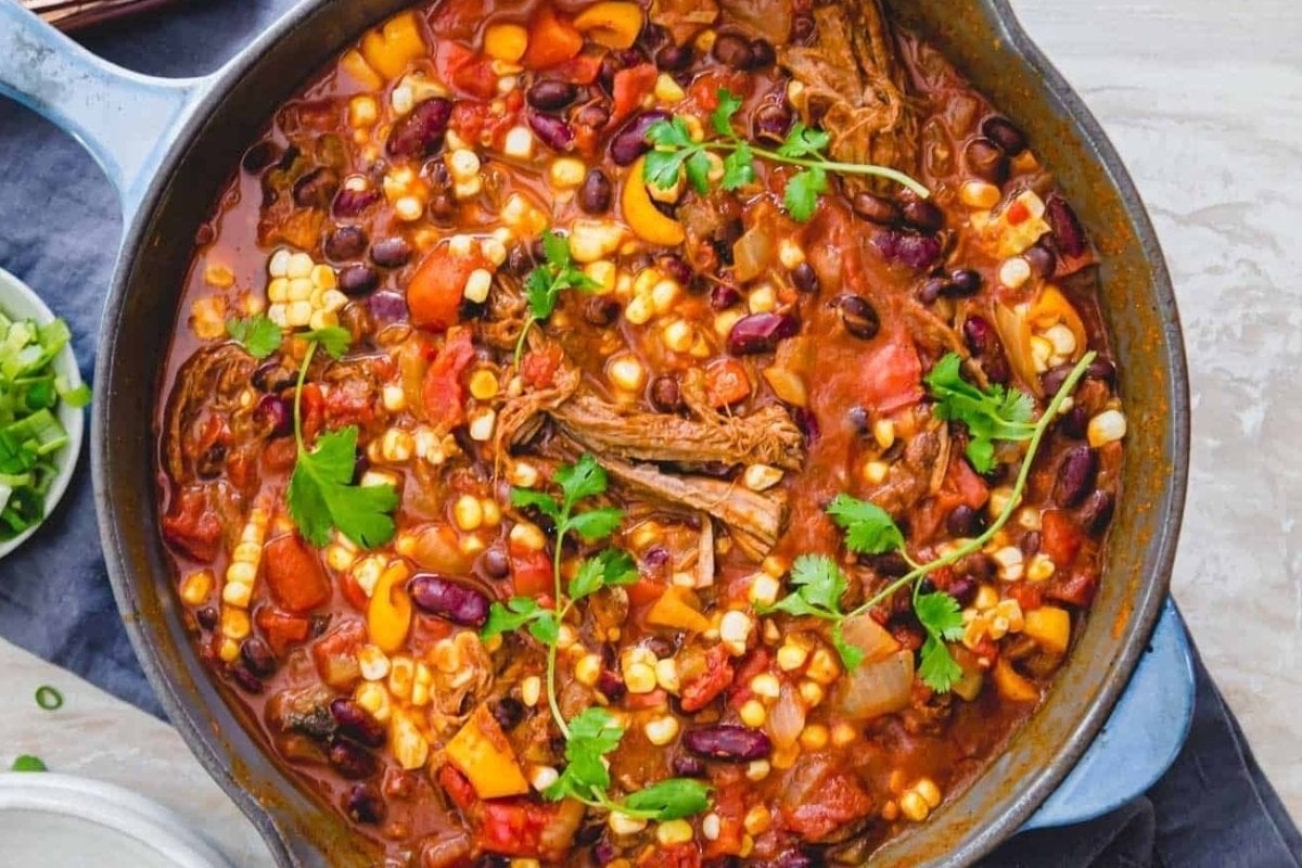 A large skillet filled with hearty chili, featuring shredded meat, beans, corn, and topped with fresh cilantro. This dish is a classic example of comfort food recipes.