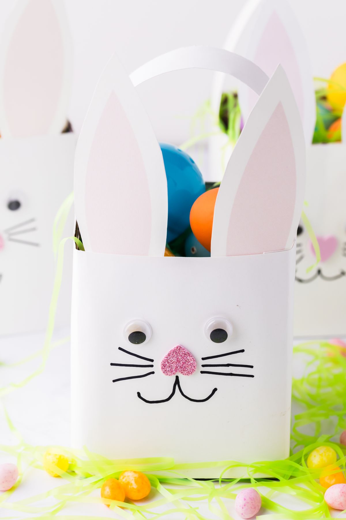 White bunny tissue box Easter basket with googly eyes, paper ears, and whiskers, filled with colorful Easter eggs.