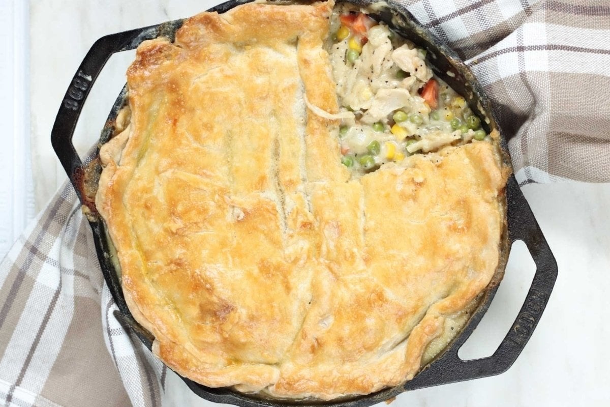 A golden-brown chicken pot pie, a classic among comfort food recipes, in a black cast iron skillet, showing a flaky crust and visible mixed vegetables inside, on a white wooden surface with a checkered napkin.