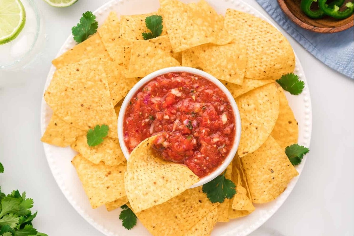 A plate of tortilla chips surrounding a bowl of salsa, garnished with cilantro on a light-colored table, perfect as bites for dinner.