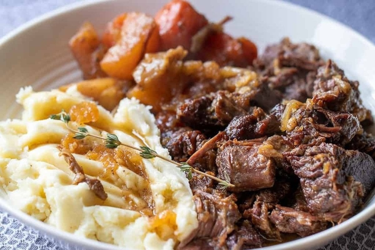 A plate of comfort food featuring mashed potatoes with gravy, pot roast, and roasted carrots and potatoes.