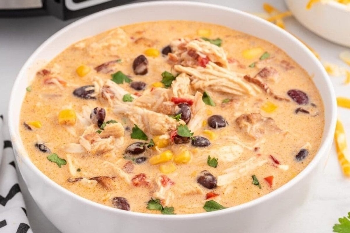 A bowl of creamy chicken soup, perfect for comfort food recipes, garnished with corn, black beans, and cilantro on a kitchen counter.