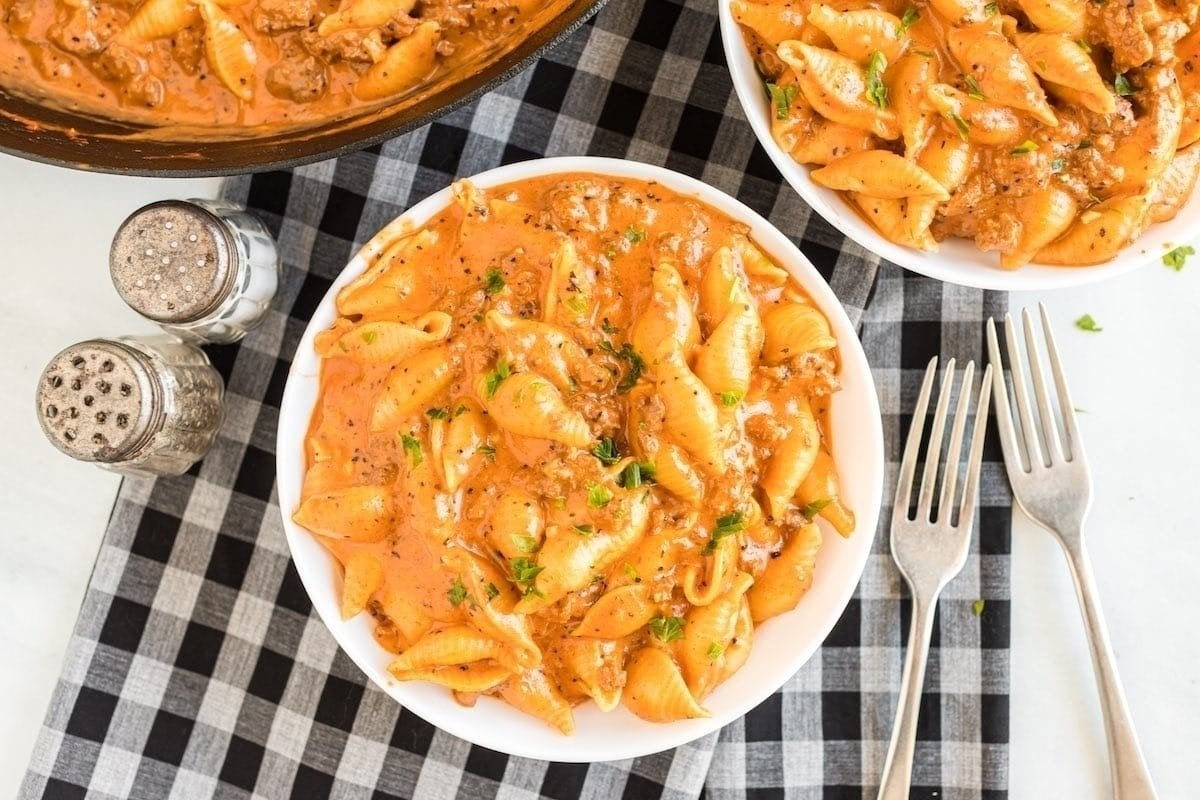 A plate of creamy tomato pasta garnished with herbs, served on a checkered tablecloth, with a skillet and additional plates of comfort food pasta recipes in the background.
