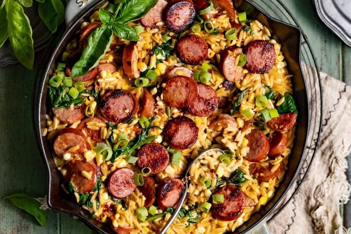 A dish of orzo pasta with sliced sausage, spinach, and scallions, garnished with basil, served in a decorative bowl as a comfort food recipe.